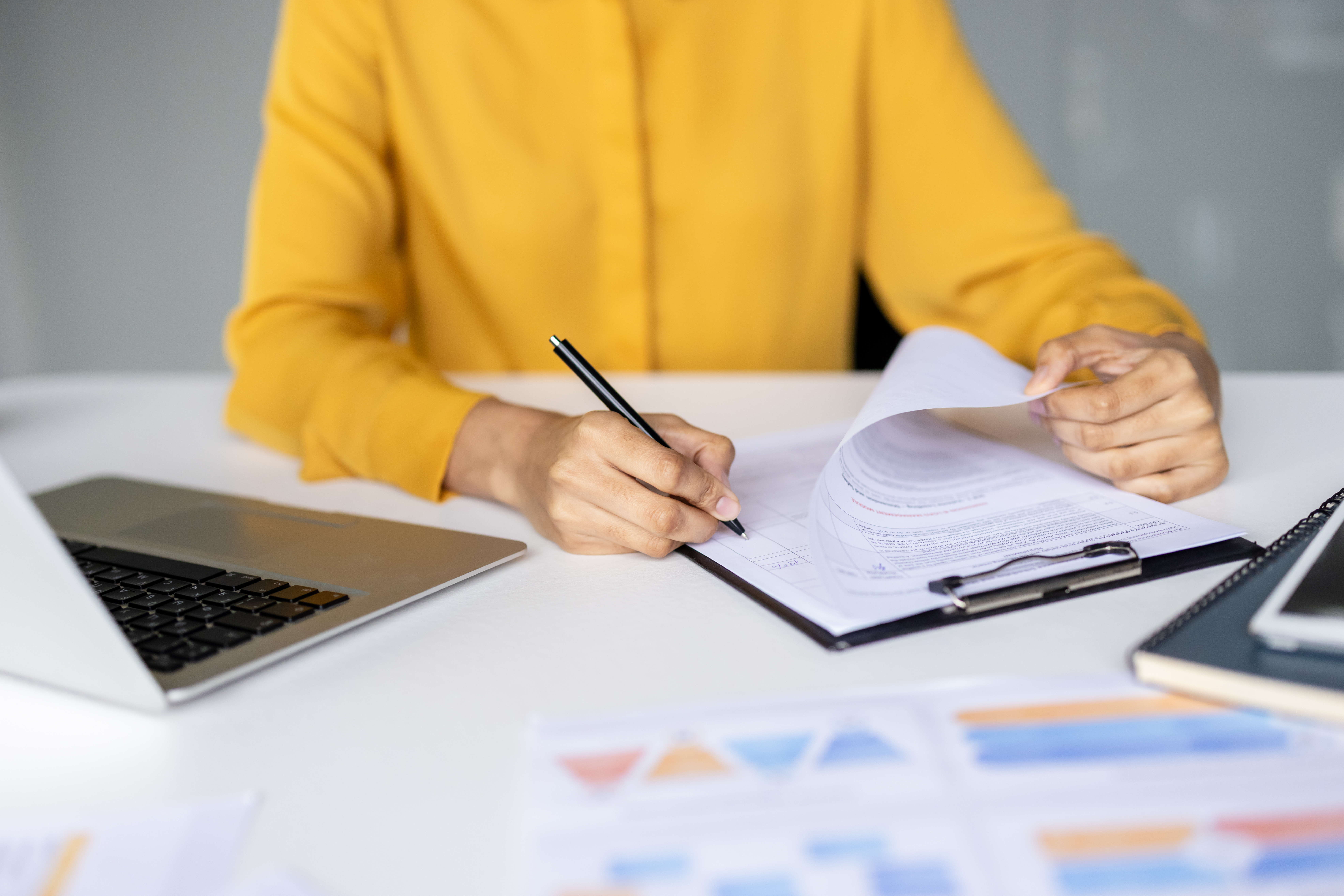 Businesswoman in a yellow shirt writing and signing documents on a clipboard, managing paperwork with a laptop and financial reports on a white desk in an office