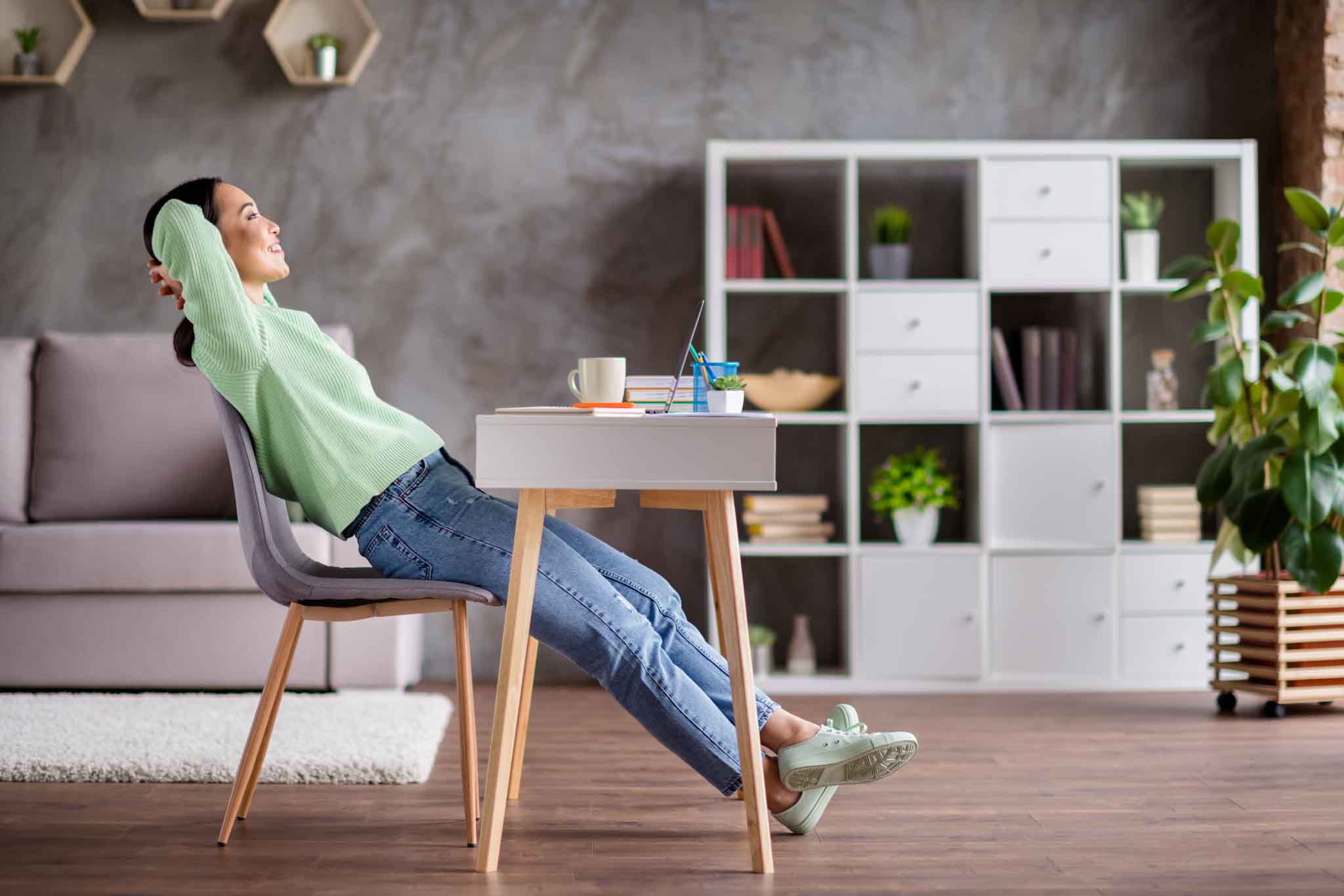 Full length profile side photo of positive korean girl have free time, end college presentation relax stretch hands wear jeans jumper sit table desk in house indoors